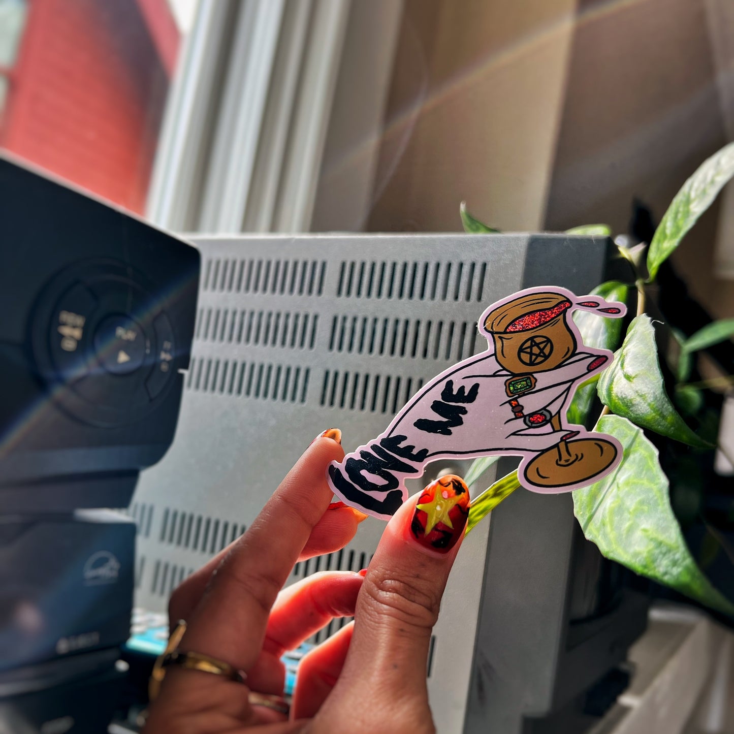 Hand holding a vinyl sticker with 'LOVE ME' text and hand pouring out a chalice.