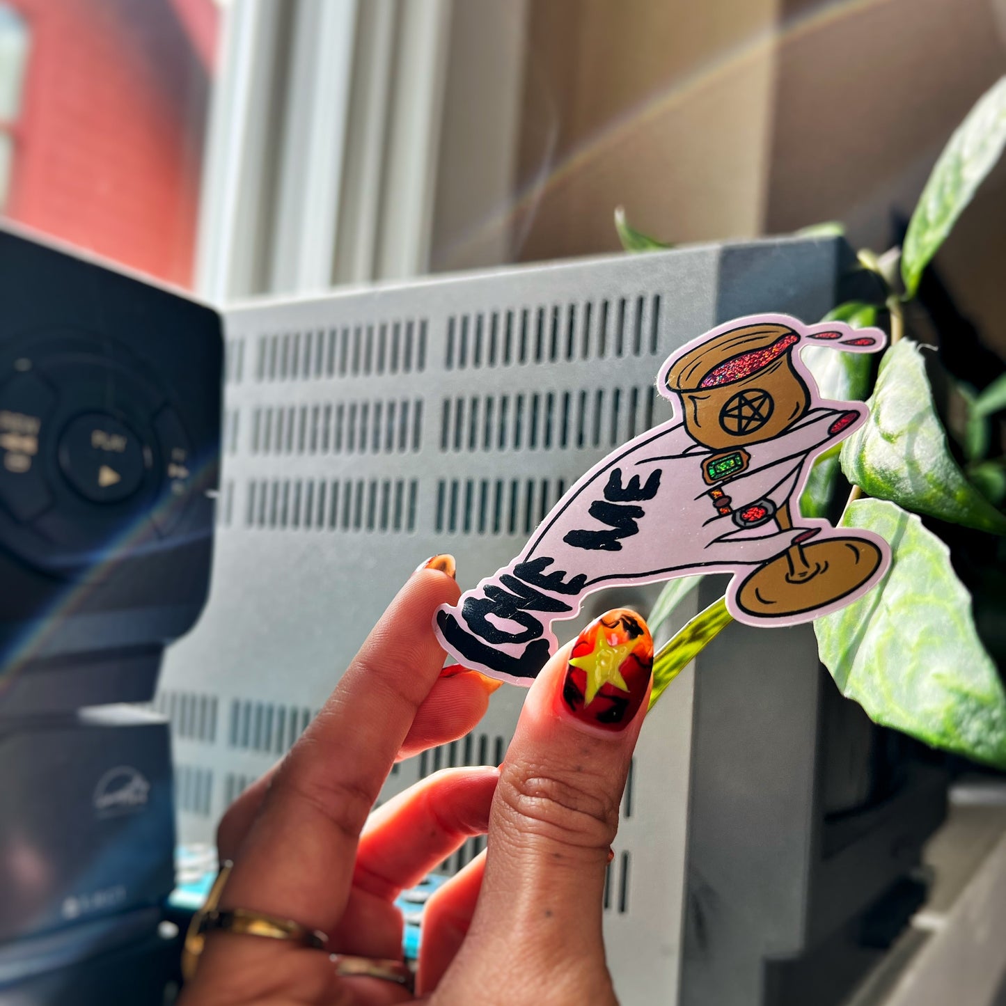 Hand holding a colorful sticker with The Love Witch (2016) inspired text and graphics, blurred indoor background