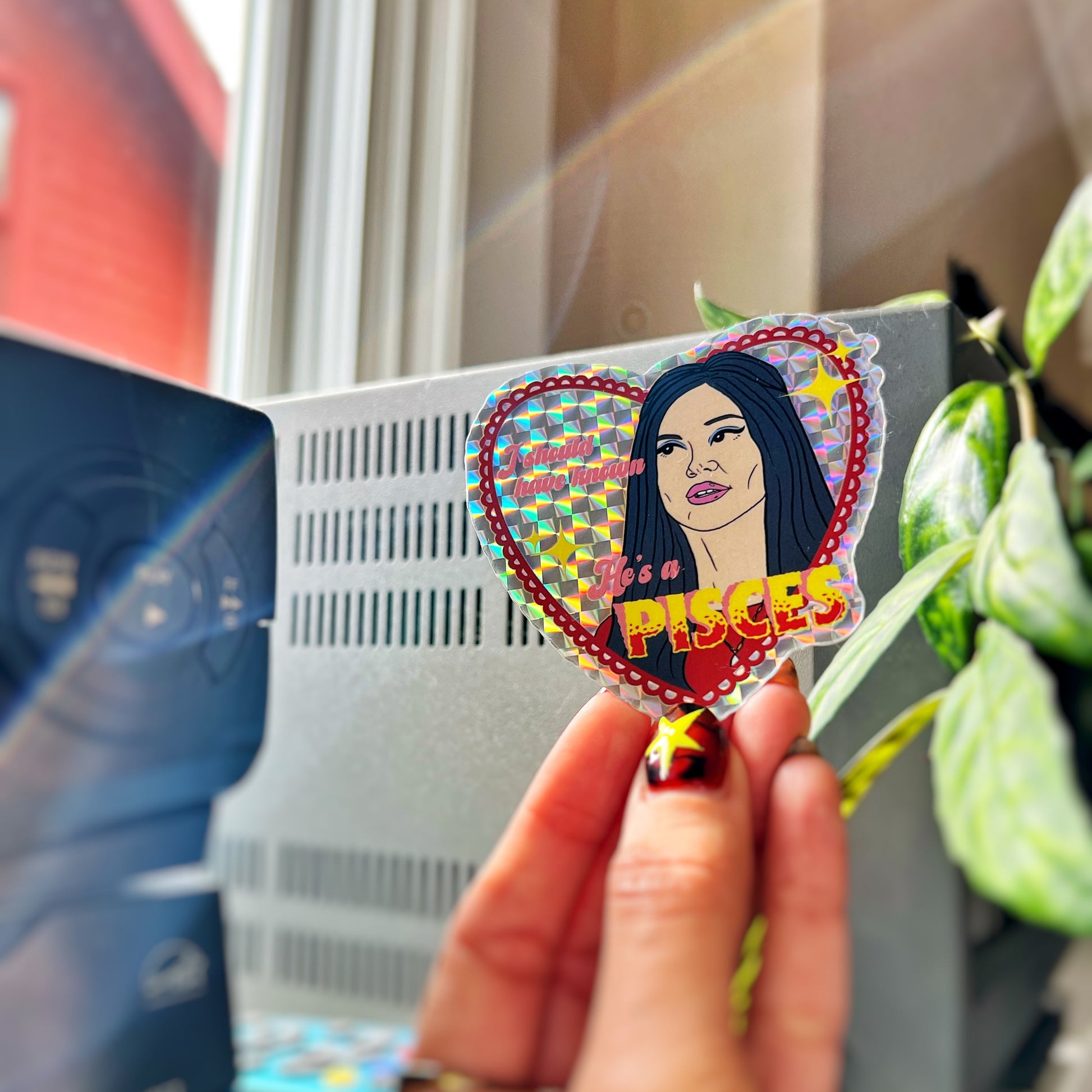 Heart-shaped sticker with a person's face and 'Pisces' text held by a hand, with a blurred background.