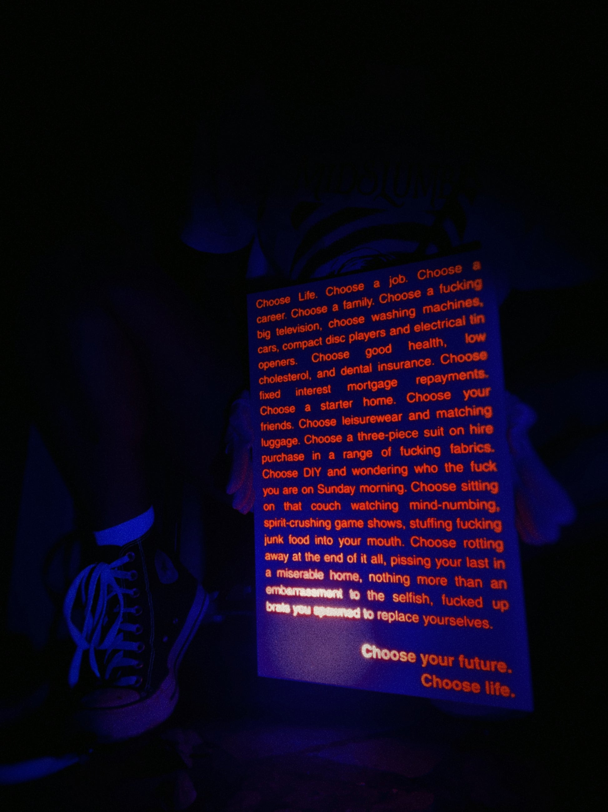 A rectangular sign with uv-light reactive orange text on a dark background, reading the opening monologue from the film Trainspotting. The sign is photographed in a dim environment with blue blacklight lighting.
