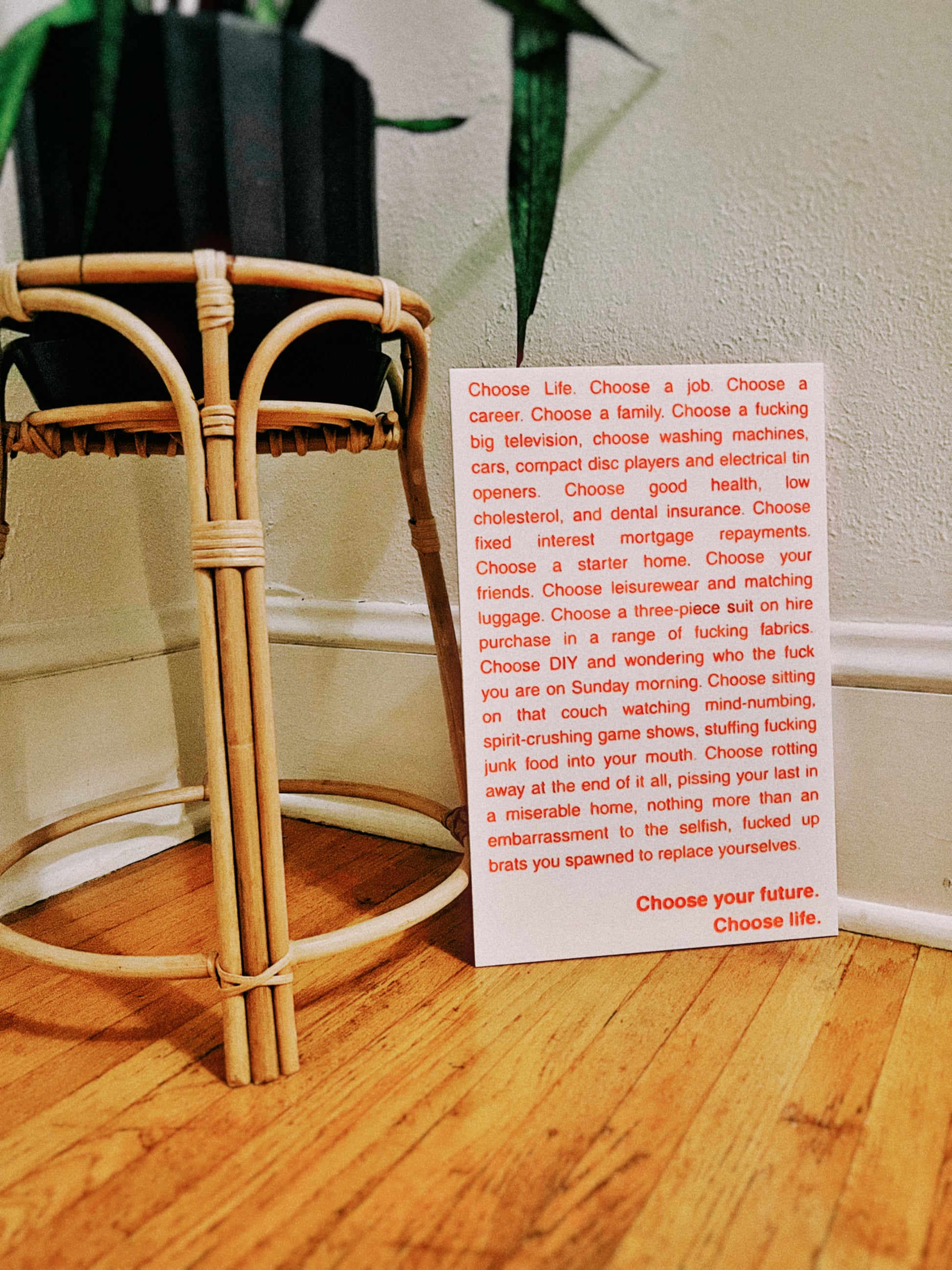 A white rectangular card with orange engraved and handpainted text that reads out the opening monologue of the film Trainspotting, placed on a wooden floor in front of a woven rattan plant stand with a potted plant and light wall in the background.