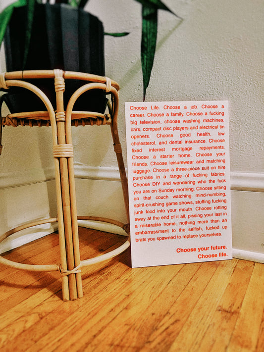 A white rectangular card with orange engraved and handpainted text that reads out the opening monologue of the film Trainspotting, placed on a wooden floor in front of a woven rattan plant stand with a potted plant and light wall in the background.