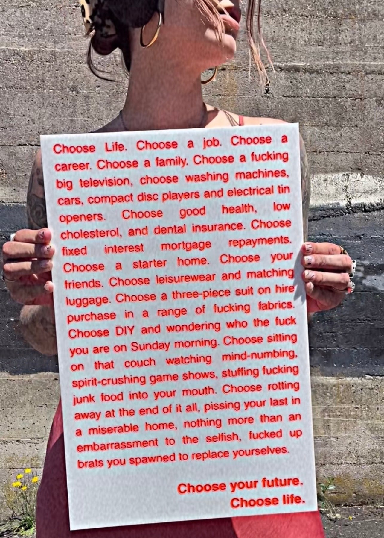 A person outdoors holding a white sign with red text that reads the opening monologue from the film Trainspotting.