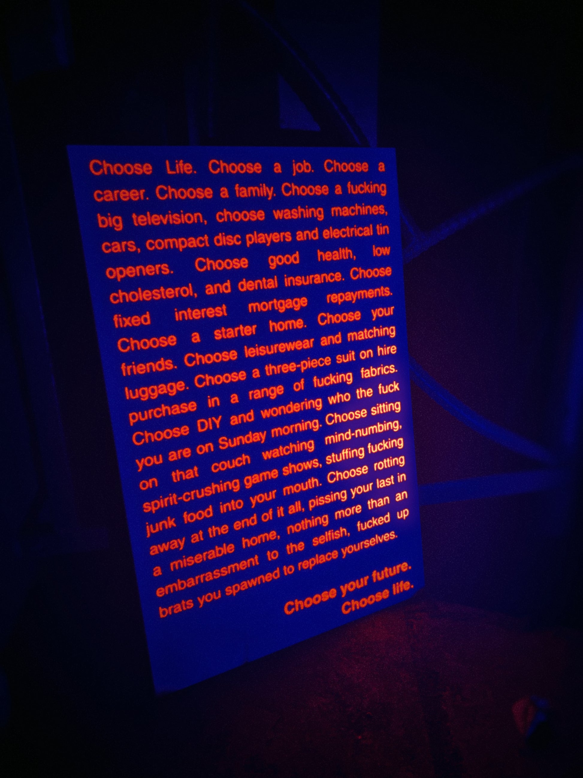 A rectangular sign with uv-light reactive orange text on a dark background, reading the opening monologue from the film Trainspotting. The sign is photographed in a dim environment with blue blacklight lighting.